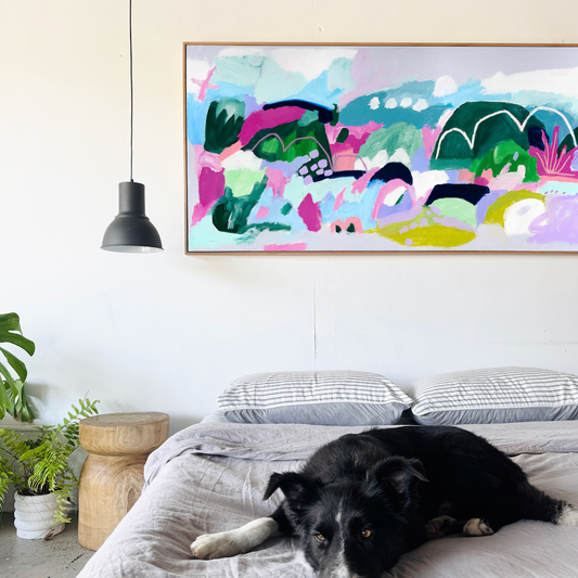 Dog lying on a bed with a colorful abstract painting above it in a room.