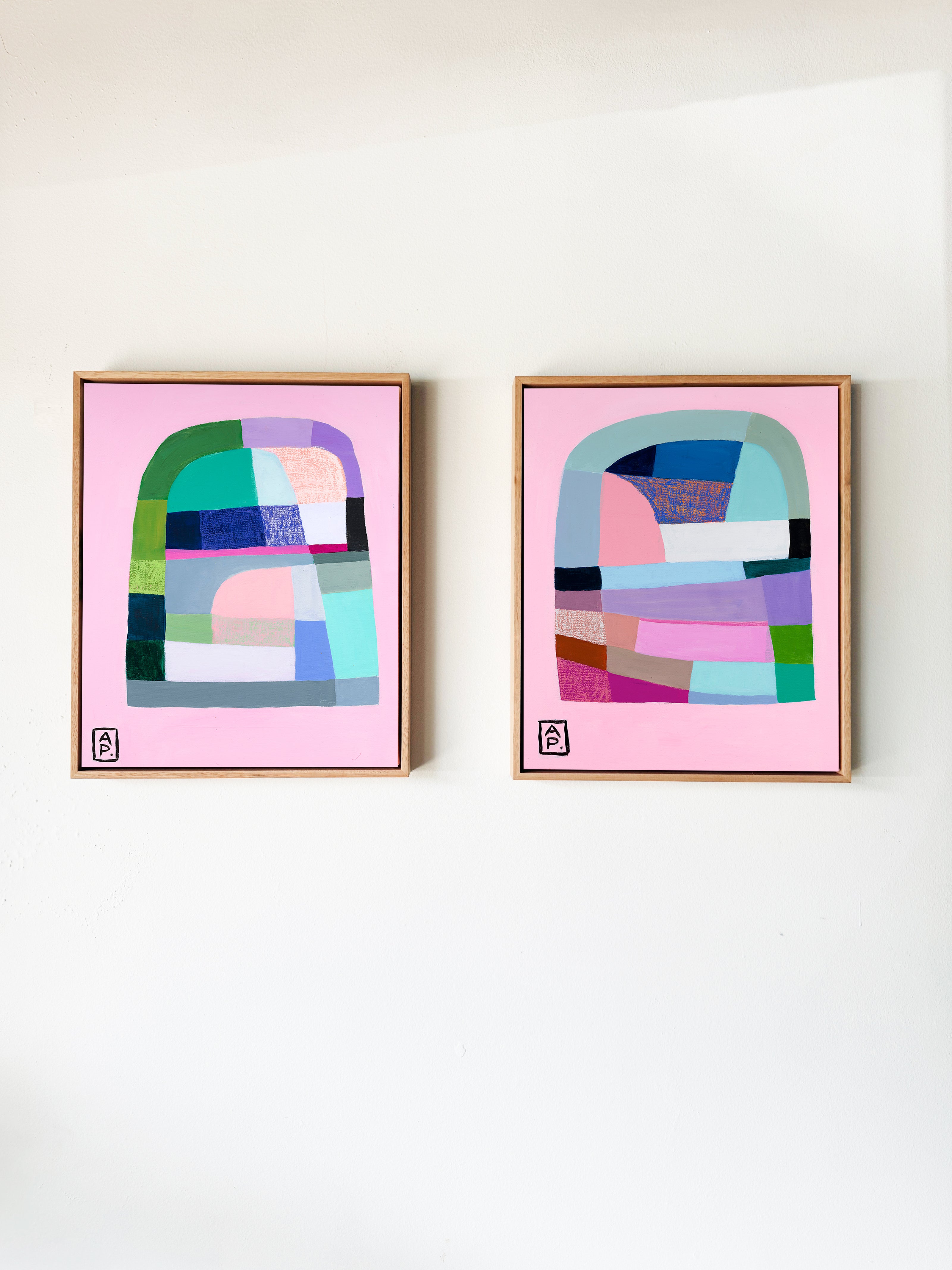 Two abstract art prints with colorful geometric designs on pink backgrounds, framed in wooden frames, against a white wall.