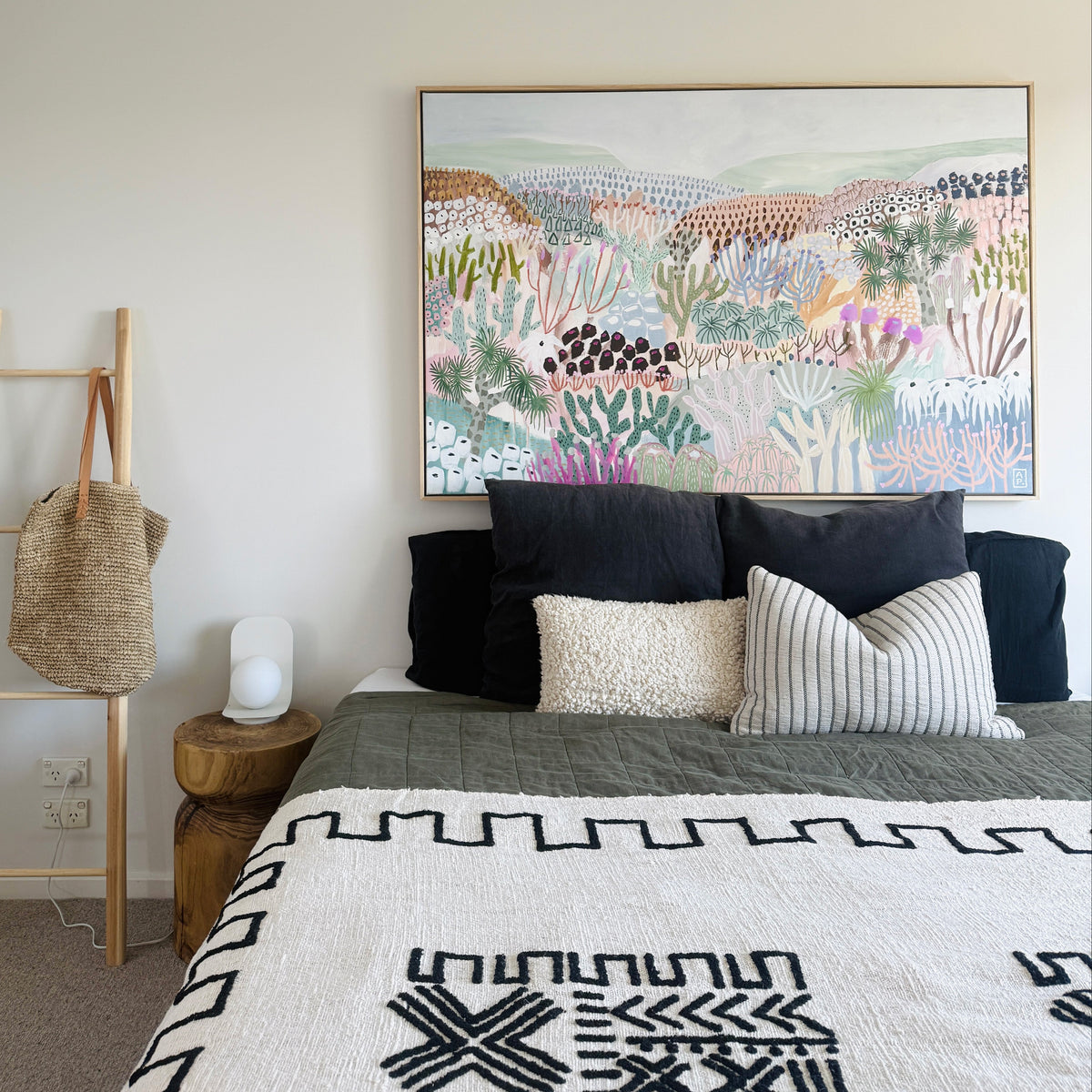 A styled bedroom scene featuring a large framed abstract landscape painting on a beige wall above a bed with black and striped pillows and a patterned white quilt. A wooden ladder shelf holding a woven bag stands to the left.
