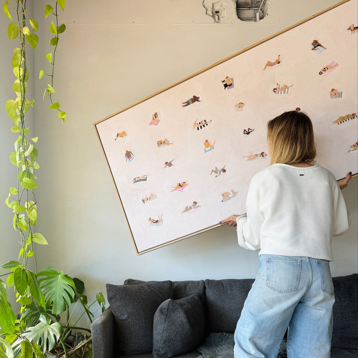 A woman stands in a living room holding a large framed art print, with a smaller matching framed print on the floor in front of a gray sofa and potted plants.
